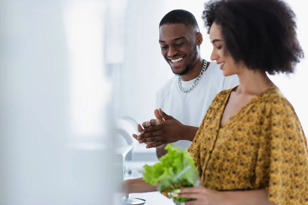 Позитивний афроамериканець, який дивиться на розмиту дівчину з салатом вдома. — стокове фото