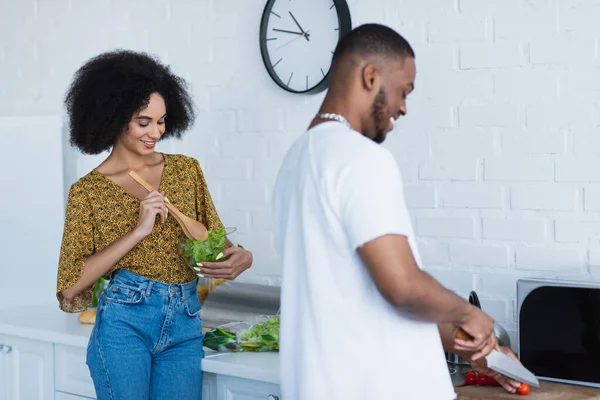 Lächelnde Afroamerikanerin mixt Salat neben Freund, der in Küche kocht — Stockfoto