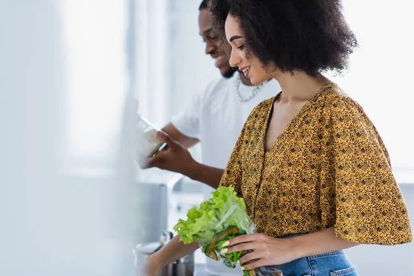 Side view of african american woman holding lettuce near blurred boyfriend at home — Stock Photo