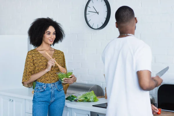 Mulher afro-americana positiva misturando salada perto namorado borrado com faca na cozinha — Fotografia de Stock