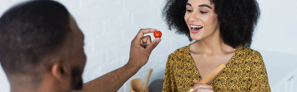 Alegre mujer afroamericana cerca borrosa novio con tomate cherry en la cocina, pancarta - foto de stock