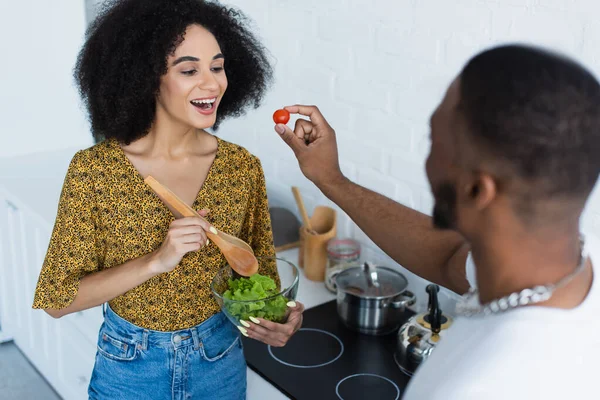 Blurred african american man feeding tomato to girlfriend in kitchen — Stock Photo