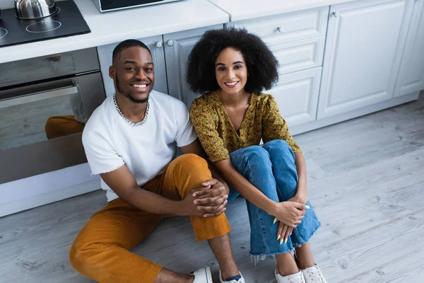 Vue grand angle du couple afro-américain souriant à la caméra sur le sol dans la cuisine — Photo de stock