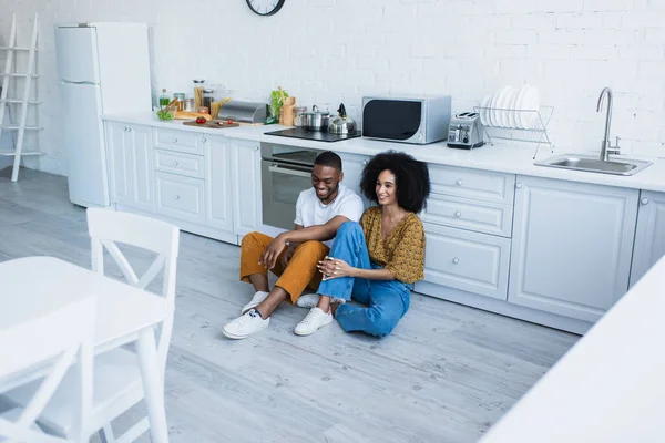 Усміхнена жінка сидить на підлозі на кухні біля афроамериканського хлопця — стокове фото