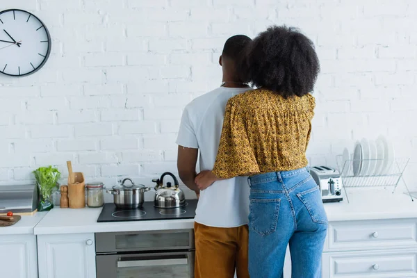 Vista posteriore della donna afro-americana che abbraccia l'uomo vicino alla stufa in cucina — Foto stock