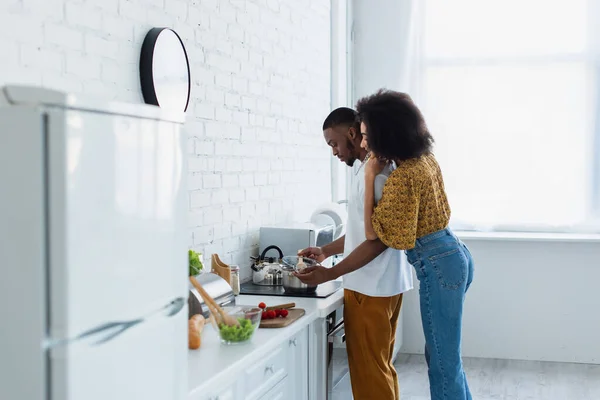 Side view of woman hugging african american boyfriend cooking in kitchen — Stock Photo