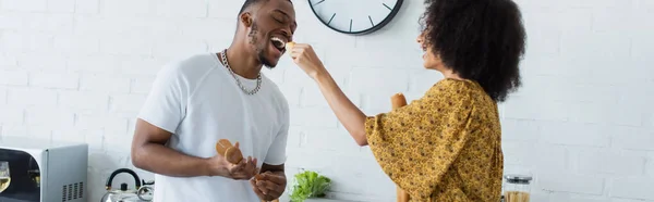 African american woman holding baguette near boyfriend with open mouth in kitchen, banner — Stock Photo
