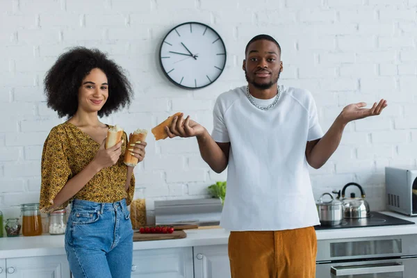 Confused african american man standing near smiling girlfriend with baguette — Stock Photo