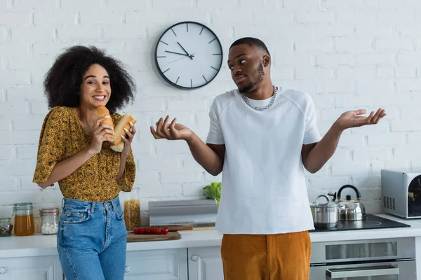 Positive african american woman holding baguette near confused boyfriend in kitchen — Stock Photo