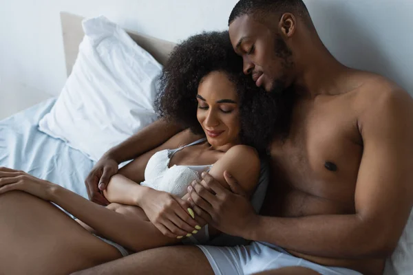 Africano americano hombre con los ojos cerrados abrazando novia en sujetador en la cama - foto de stock