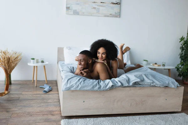 Sonriente mujer afroamericana en lencería acostada en su novio en la cama - foto de stock