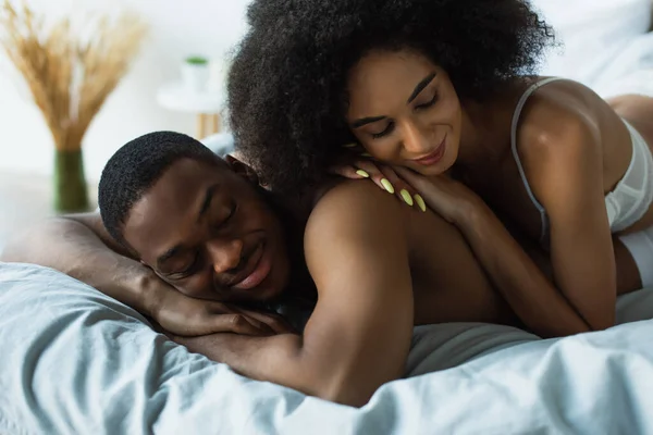 Afroamerikanerin im BH liegt auf Freund mit geschlossenen Augen auf Bett — Stockfoto