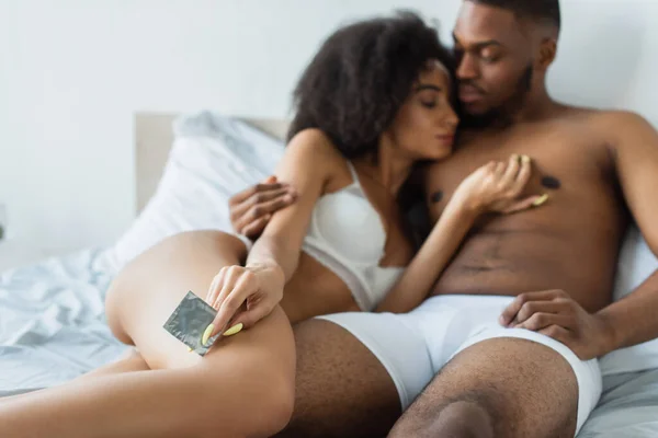 Презерватив в руке размытой африканской женщины рядом с парнем на кровати — стоковое фото