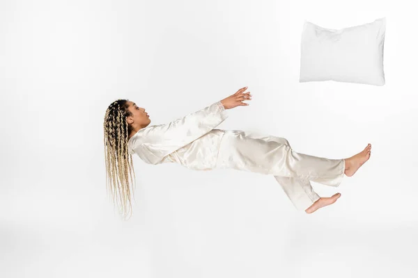 Pillow flying away from african american woman levitating in silk pajamas isolated on white — Stock Photo