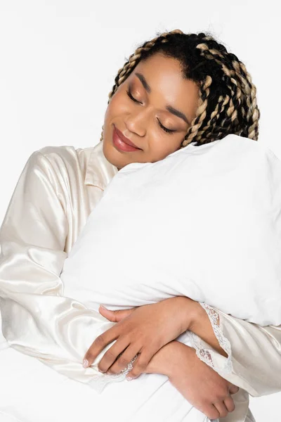 Schläfrige afrikanisch-amerikanische Frau lächelt mit geschlossenen Augen, während sie Kissen isoliert auf weiß umarmt — Stockfoto