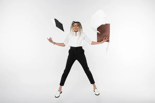 Alegre mulher de negócios afro-americana levitando perto de pastas e documentos isolados em branco — Fotografia de Stock