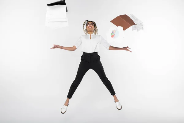 Emocionada mujer de negocios afroamericana levitando cerca de carpetas y documentos con gráficos aislados en blanco - foto de stock