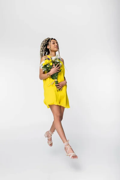Positive african american woman in yellow dress holding flowers while walking in air on white — Stock Photo