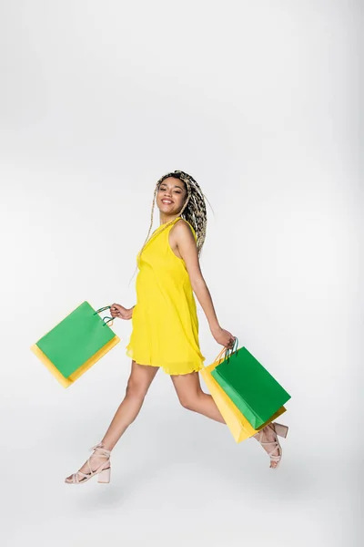 Alegre mujer afroamericana sonriendo a la cámara mientras levita con bolsas de compras en blanco - foto de stock