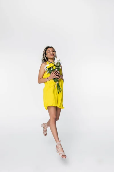 Alegre mujer afroamericana en vestido amarillo brillante caminando en el aire con flores en blanco - foto de stock