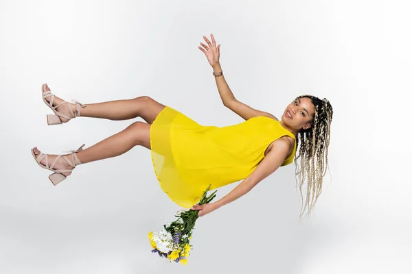 Mujer afroamericana en vestido amarillo levitando con ramo aislado en blanco - foto de stock