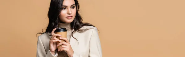 Joven bonita mujer en cuello alto sosteniendo taza de papel y mirando hacia otro lado aislado en beige, bandera — Stock Photo