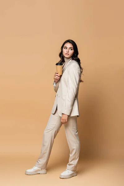 Full length of young woman in turtleneck and suit holding coffee to go on beige — Stock Photo