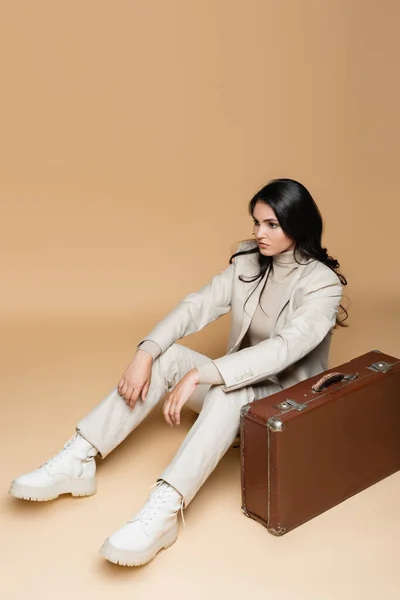 Young woman in suit sitting near vintage suitcase on beige — Stock Photo