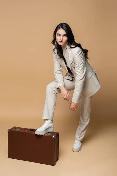 Повна довжина молодої моделі в костюмі позує, стоячи біля старовинної валізи на бежевому — стокове фото