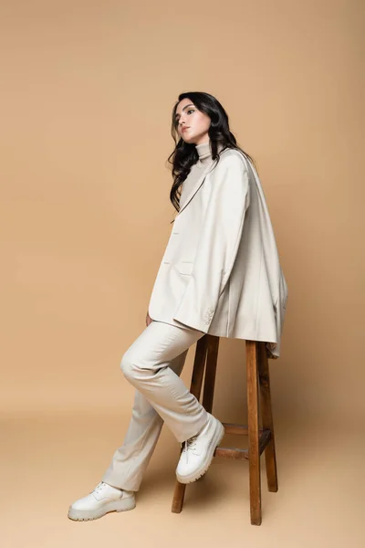 Pleine longueur de jeune femme en costume posant tout en étant assis sur tabouret en bois sur beige — Photo de stock
