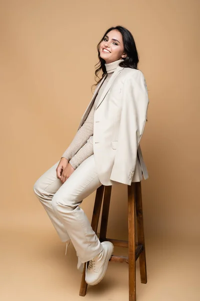 Full length of cheerful woman in suit posing while sitting on wooden stool on beige — Stock Photo