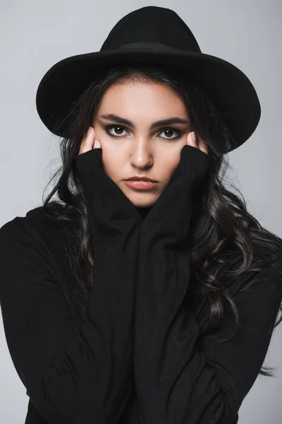 Молодая модель в черной шляпе и водолазке, смотрящая на камеру, изолированную на сером — стоковое фото