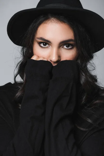 Young model in black fedora hat and turtleneck looking at camera while covering mouth isolated on grey — Stock Photo