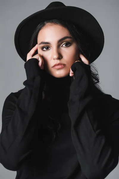 Brunette young model in black fedora hat and turtleneck looking at camera isolated on grey — Stock Photo