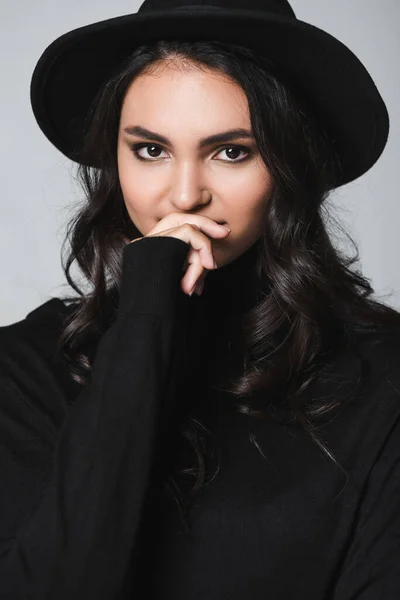 Model in black fedora hat and turtleneck covering mouth while looking at camera isolated on grey — Stock Photo