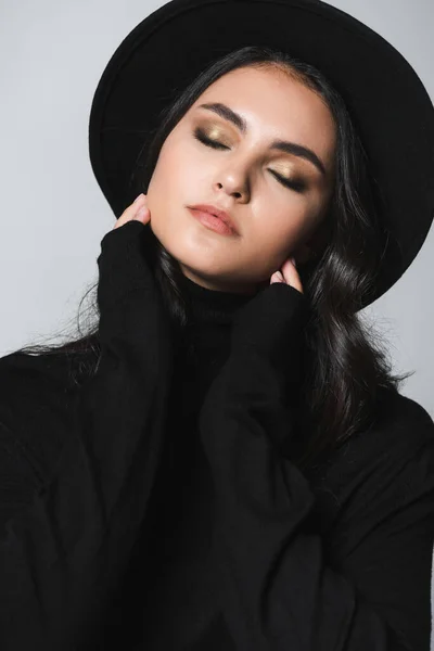 Молодая модель с закрытыми глазами в черной шляпе fedora и водолазке изолированы на сером — стоковое фото