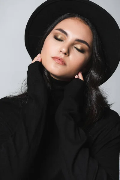 Modelo joven con ojos cerrados en sombrero fedora negro y cuello alto aislado en gris - foto de stock