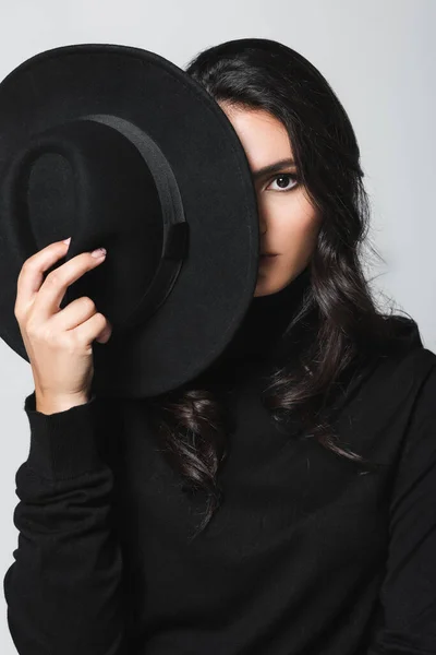 Молодая модель, держащая черную шляпу Fedora, закрывая лицо и глядя на камеру, изолированную на сером — стоковое фото