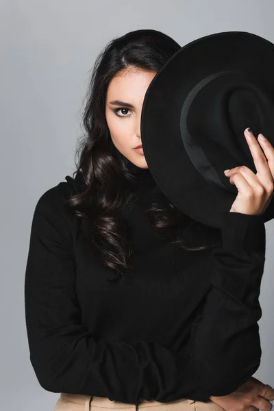 Mujer joven sosteniendo sombrero fedora negro mientras cubre la cara y mirando a la cámara aislada en gris - foto de stock