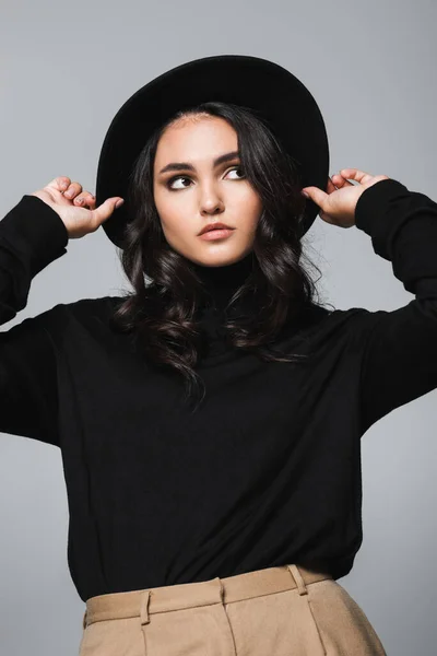 Brunette et jeune modèle ajustant chapeau fedora noir et regardant loin isolé sur gris — Photo de stock