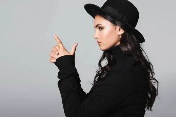 Vista lateral de morena y joven modelo en sombrero fedora mostrando gesto de pistola de dedo aislado en gris - foto de stock