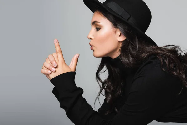 Side view of brunette model in fedora hat pouting lips and showing finger gun gesture isolated on grey — Stock Photo