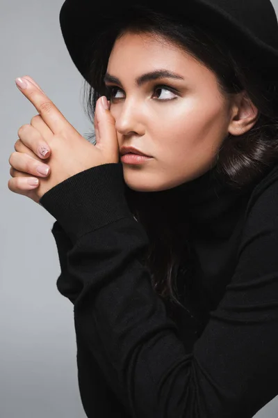 Junge brünette Frau mit Fedora-Hut schaut weg und zeigt Fingergewehr Geste isoliert auf grau — Stockfoto