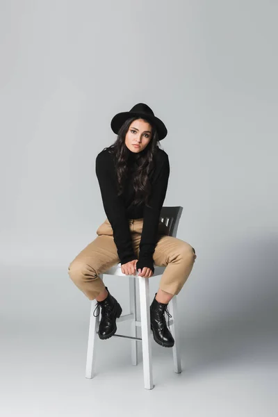 Volle Länge der jungen und trendigen Frau mit Fedora-Hut, die in die Kamera schaut, während sie auf einem Stuhl auf grau sitzt — Stockfoto