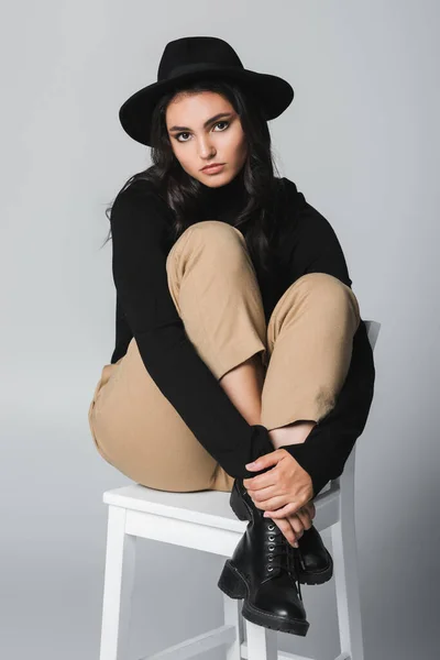 Full length of brunette model in fedora hat looking at camera while sitting on chair on grey — Stock Photo