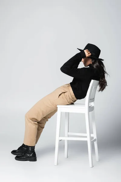 Side view of young brunette woman in fedora hat leaning on chair on grey — Stock Photo
