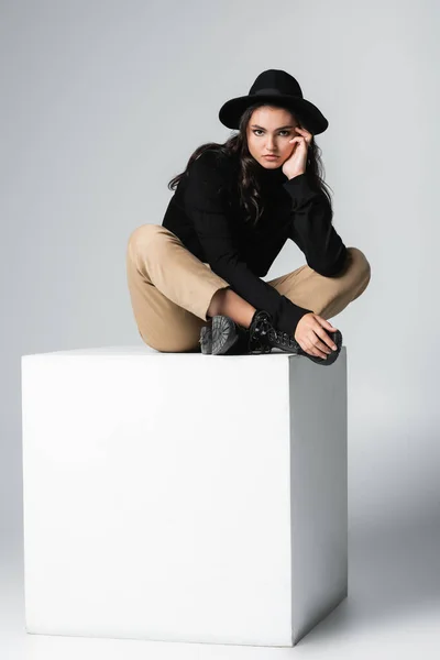 Full length of stylish model in fedora hat looking at camera while sitting on white cube on grey — Stock Photo