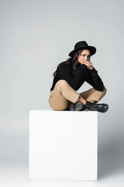 Longueur totale du modèle onéreux en chapeau fedora assis avec jambes croisées sur cube blanc sur gris — Photo de stock