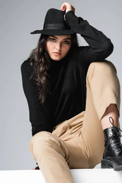 Stylisches Modell mit Fedora-Hut, das in die Kamera schaut, während es auf einem weißen Würfel sitzt, isoliert auf grau — Stockfoto