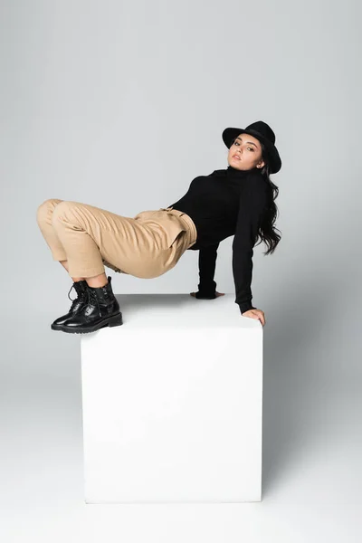 Toute la longueur du modèle élégant en chapeau fedora regardant la caméra tout en posant sur le cube blanc sur gris — Photo de stock